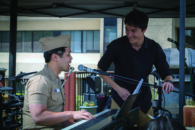 A man in brown Navy uniform plays the piano and wears a harmonica on a neckstrap while another man looks on