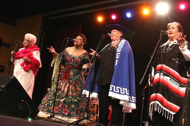 Four women dressed in Maexican attire stand before microphones on a stage.