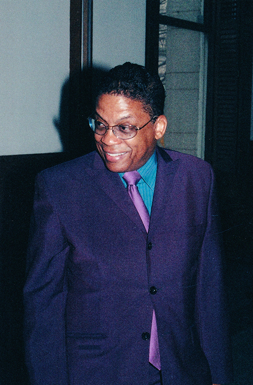 Man smiling wearing glasses and a purple suit.