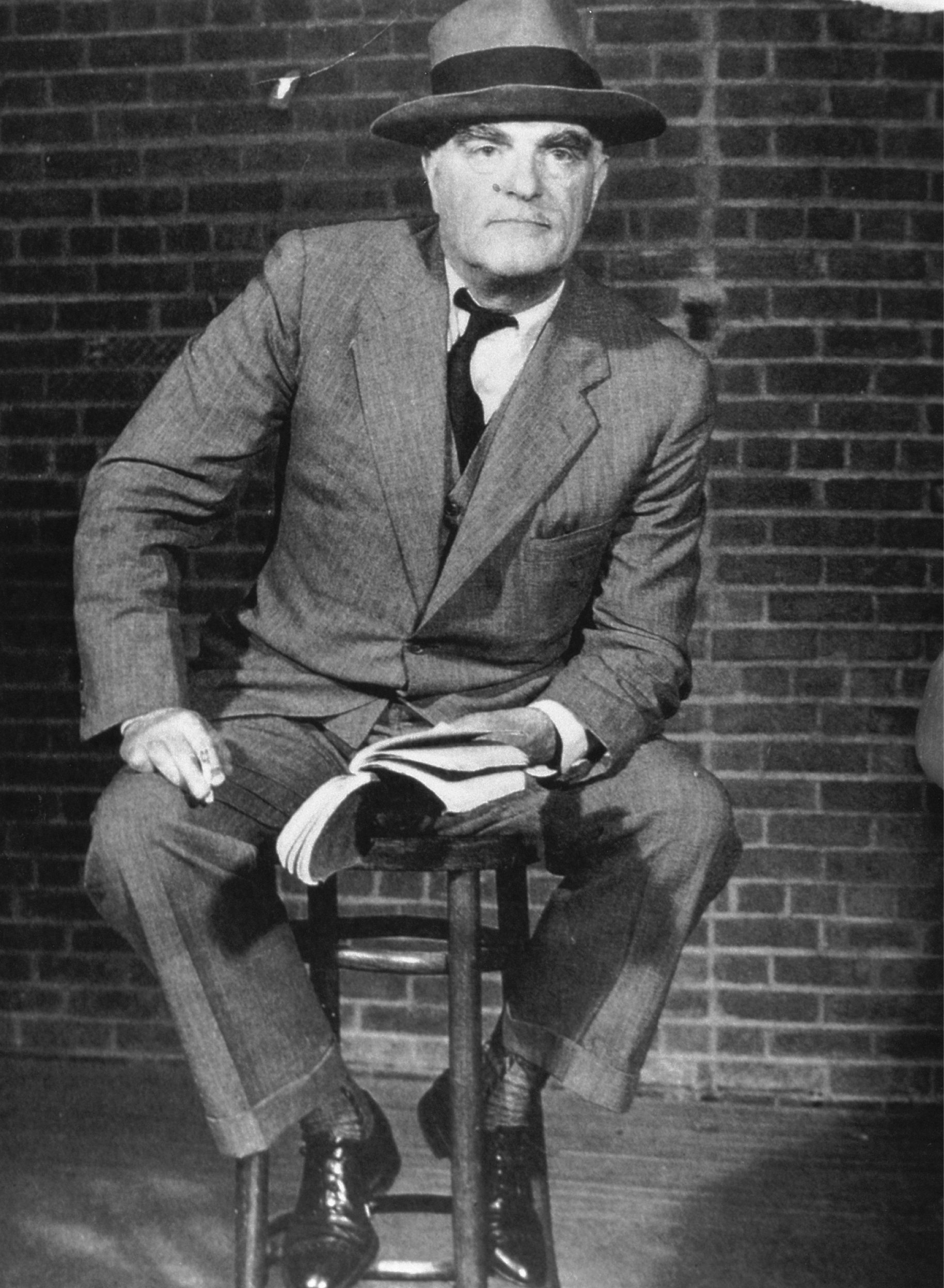 Man in suit and hat sitting on barstool holding a book on stage. 