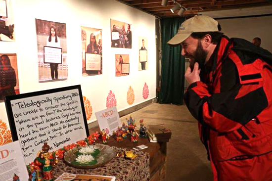 Man reads a quote about “ABCD Life” written on a whiteboard. Whiteboard rests on a table decorated with Hindu statues, flowers, and copies of The Namesake. Behind, a wall displays photographs as part of the exhibit. 