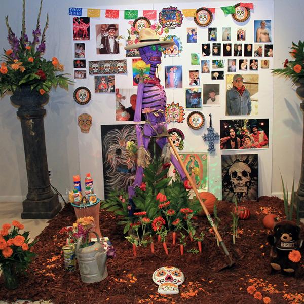 An altar for a Day of the Dead celebration with a purple skeleton, orange flowers, and a poster with photos on it. 