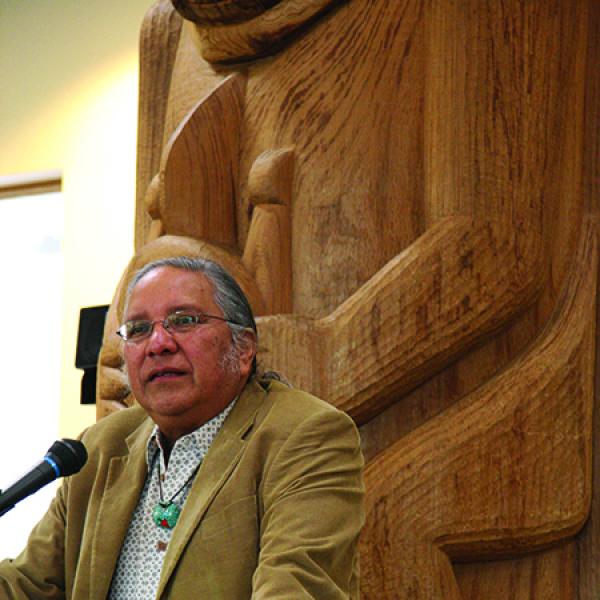 Native-American man at podium in front of large wooden carving. 