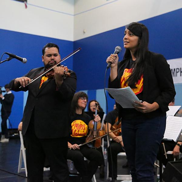 A man playing a violin stands next to a woman speaking into a microphone