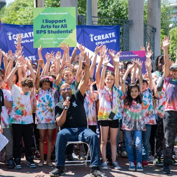  A group of children celebrate the arts in Boston Public Schools. They are outside with their hands raised high, some holding signs that say "iCreate" and "Creative Mind at Work." Many are wearing tie-dyed t-shirts and some hold musical instruments. 