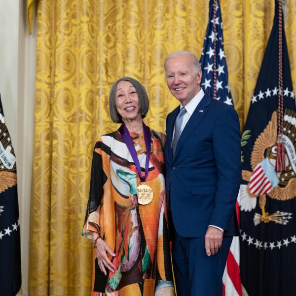 Older white male in blue suit posing with older Asian woman in colorful dress in front of flags and gold curtain.