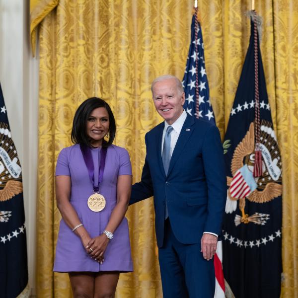 Older white male in blue suit posing with Indian woman in purple dress in front of flags and gold curtain.