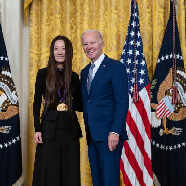 Older white male in blue suit posing with Asian woman in black dress in front of flags and gold curtain.