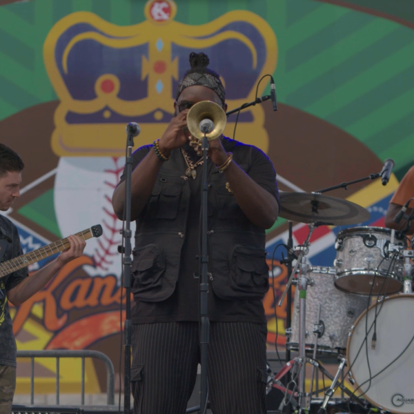 On a music stage: Black man (center) playing a trumpet, White man (left) playing the guitar, and Black man (right) playing drums.