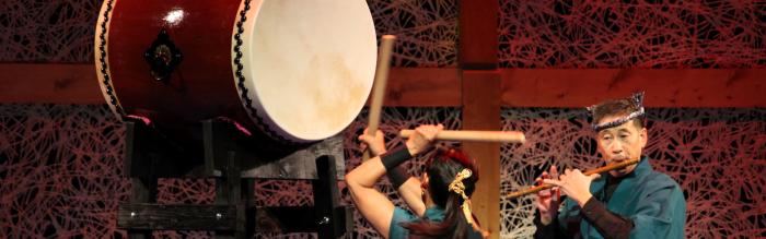 Woman banging a large drum while man plays flute on stage. 