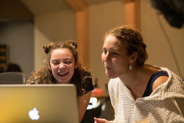 Two women laugh, looking at a laptop screen in a recording studio.
