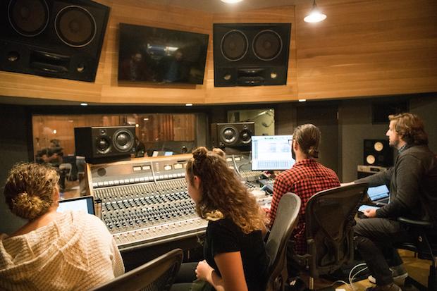 Two women and two men sit at a large mixing board in a recording studio, working.