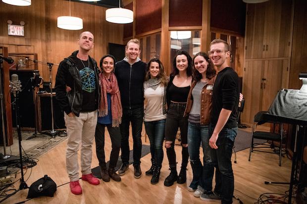 Four women and three men stand center frame in a recording studio, arms around each other, smiling.