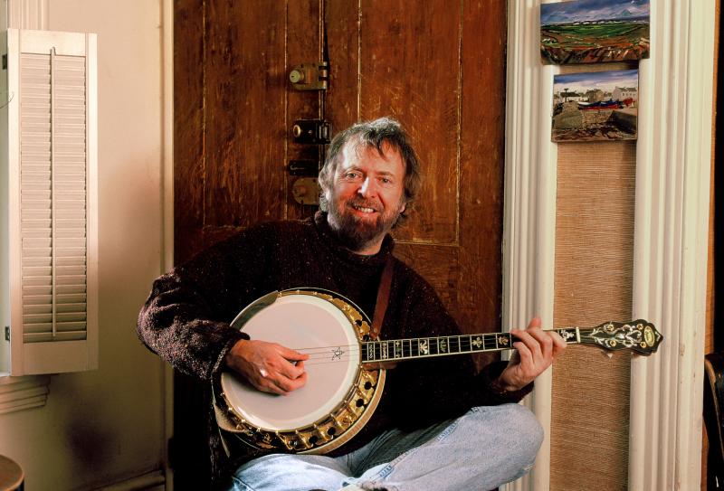 A man with brown hair and a beard smiles while strumming his tenor banjo