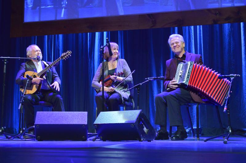 Three people play on stage with a blue background. A man with a banjo and woman with a fiddle look at a man playing the button accordian