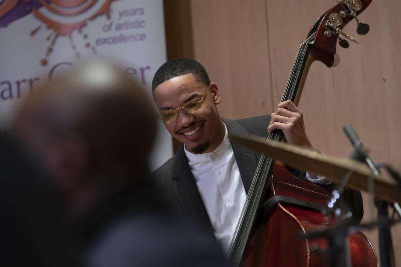 Black man wearing glasses and a black suit, playing a double bass. 