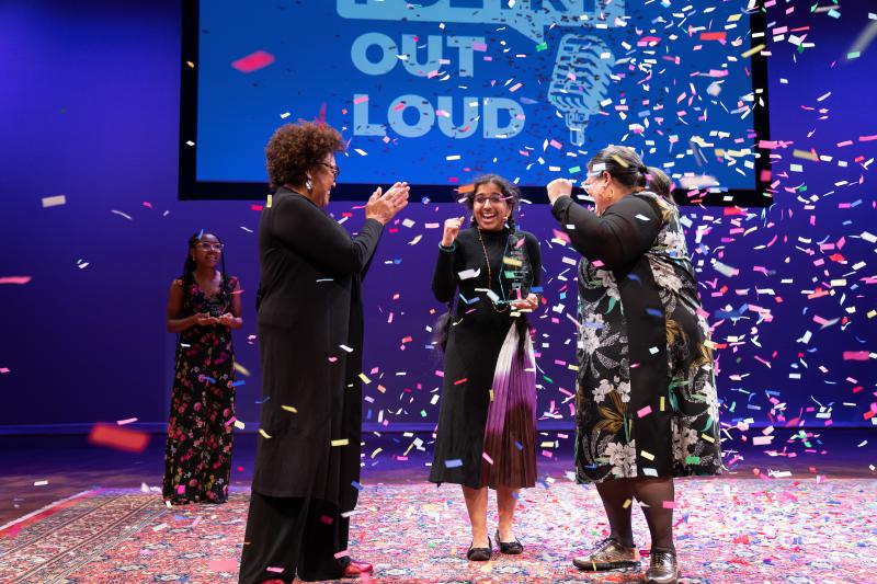 Two women of color clap on either side of a younger South Asian woman as confetti falls around them. 