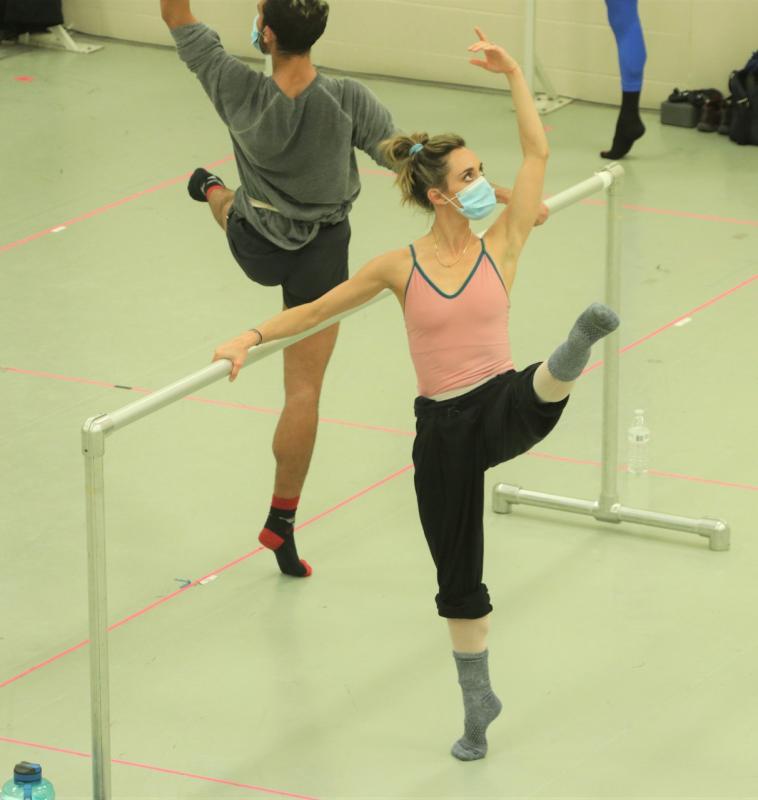 Two ballet dancers wearing masks lift their legs while rehearsing at the barre