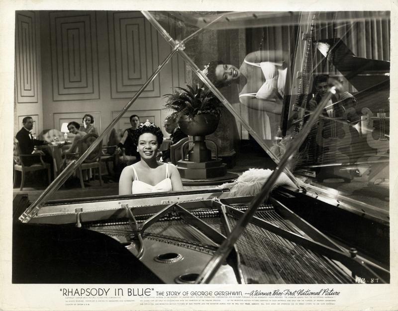 Photo of a Black woman smiling and seated at a piano with a tiara on her head, with three White women and two White men seated in the audience and watching the Black woman perform at the piano. The bottom part of the photo says: “Rhapsody in Blue” THE STORY OF GEORGE GERSHWIN ~A Warner Bros First National Picture.