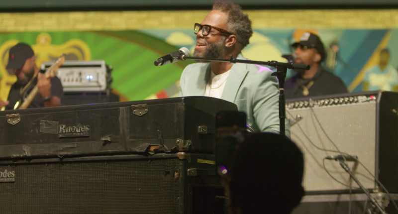 On a music stage: Black man (front and center) playing on a keyboard and singing, a Black man (left) playing the guitar, and Black man (right) singing backup to the lead vocalist.
