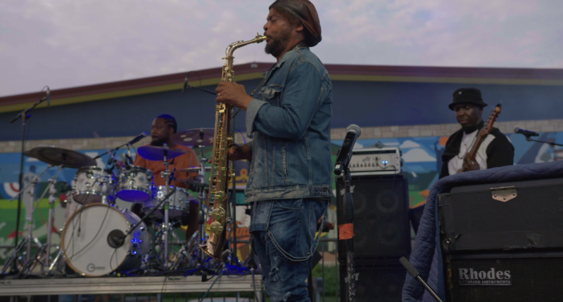 On a music stage: Black man (front and center) playing a saxophone, Black man (left) playing drums, and Black man (right) playing the guitar.