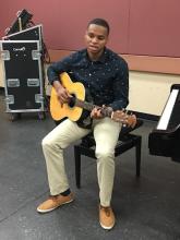 an African American young man sits on a piano stool holding his acoustic guitar
