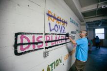 In a vacant space, artists spraypaint colorful text on a white cinderblock wall. Words include racism (crossed out), love, displacement crossed out, chain stores, homelessness, affordable rent, and gentrification.