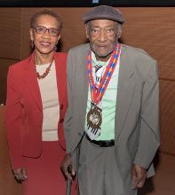 A man in a gray suit wearing a gold medal with a red, blue, and yellow ribbon stands next to a woman in a red suit.