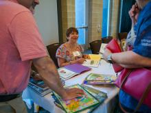 a woman sits at a conference table covered with children's book signing a copy of a book while a couple of adults look on