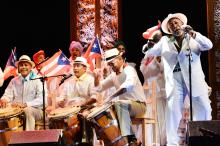 On stage: Puerto Rican man (right) wears a white hat and suit performing on stage alongside seven musicians (three men are playing drums and four women are behind them holding Puerto Rico flags).