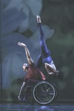 Dancer on left in red tshirt and black pants, in a wheelcair; supporting dancert on right wearing a blue leotard.