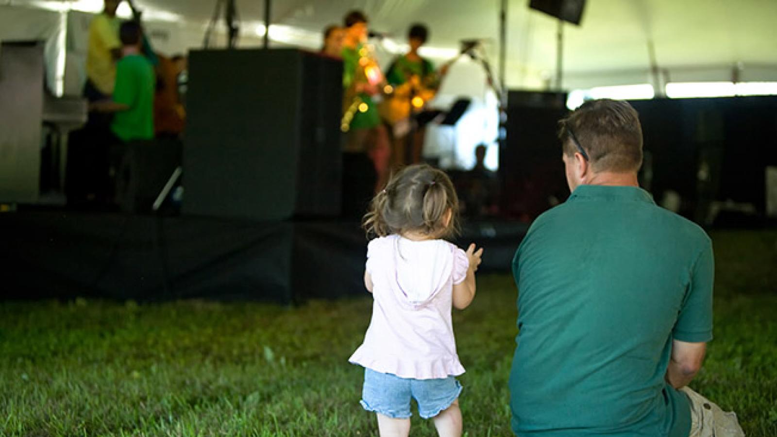A toddler girl standing in the grass next to her sitting father as they watch an outdoor performance