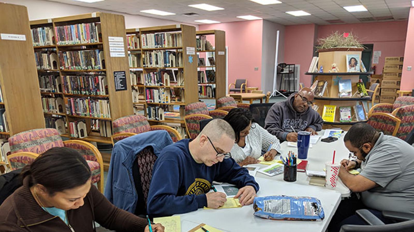 With library stacks in the background, men and women sit around a table and write on yellow legal pads