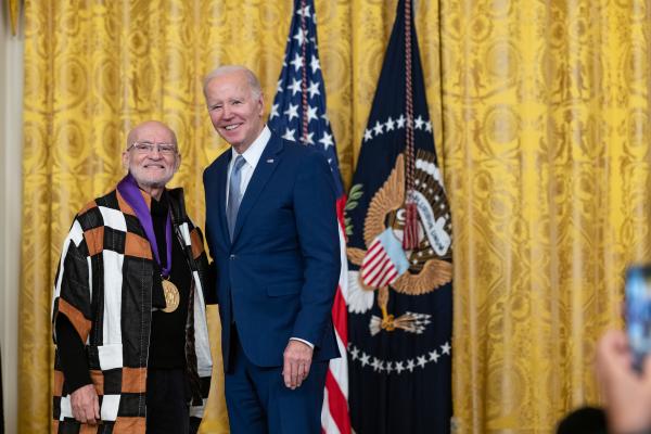 Older white male in blue suit posing with older Hispanic man wearing a black-brown-white patchwork jacket in front of flags and gold curtain.