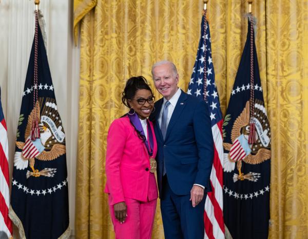 Older white male in blue suit posing with Black woman in pink outfit in front of flags and gold curtain.